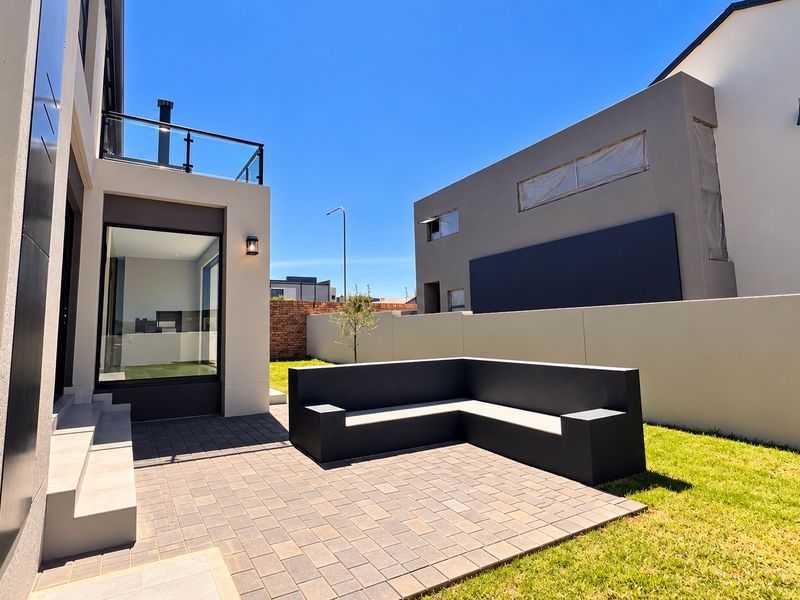 Modern rear exterior featuring a paved patio area with built-in masonry seating, a landscaped garden, and an upper-level balcony with glass balustrades.