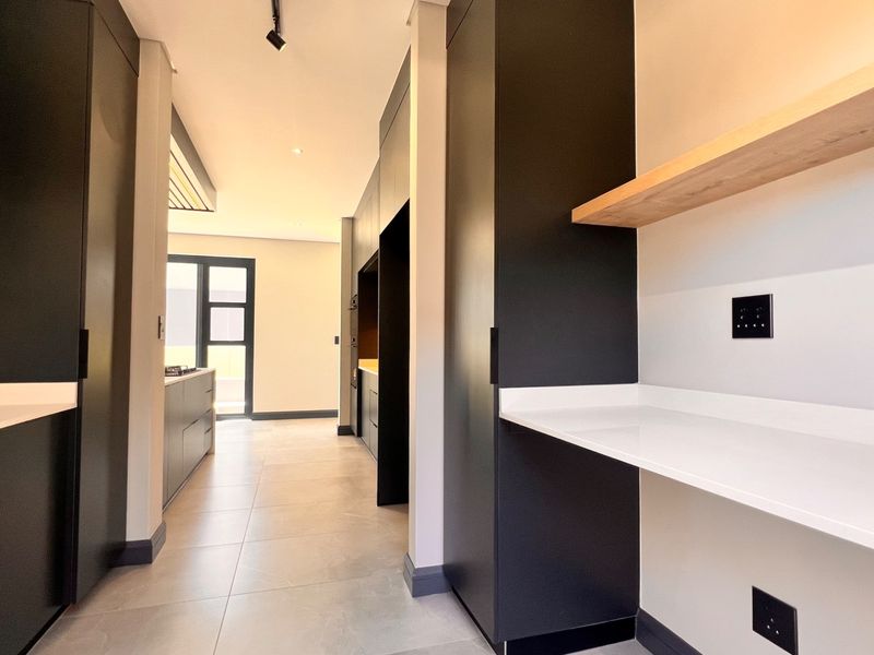 A modern, minimalist scullery featuring sleek dark cabinetry, white stone countertops, and a floating timber shelf, providing a clean and functional prep area.