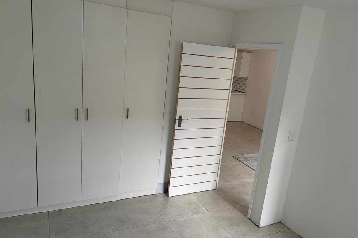 A clean, empty bedroom featuring built-in cupboards, neutral tiled flooring, and a view into the adjacent kitchen area.