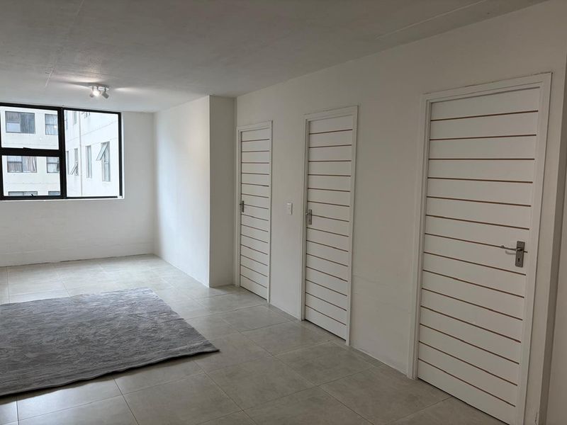 A bright, open-plan living area featuring neutral tiled flooring, clean white walls, and three internal doors leading to other rooms.