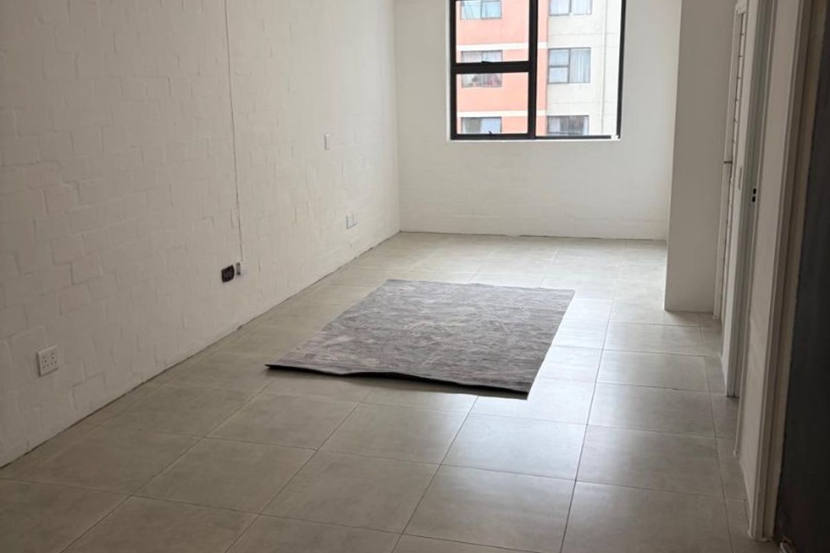 A bright, open-plan living area featuring neutral tiled flooring, painted brick walls, and large windows providing natural light.