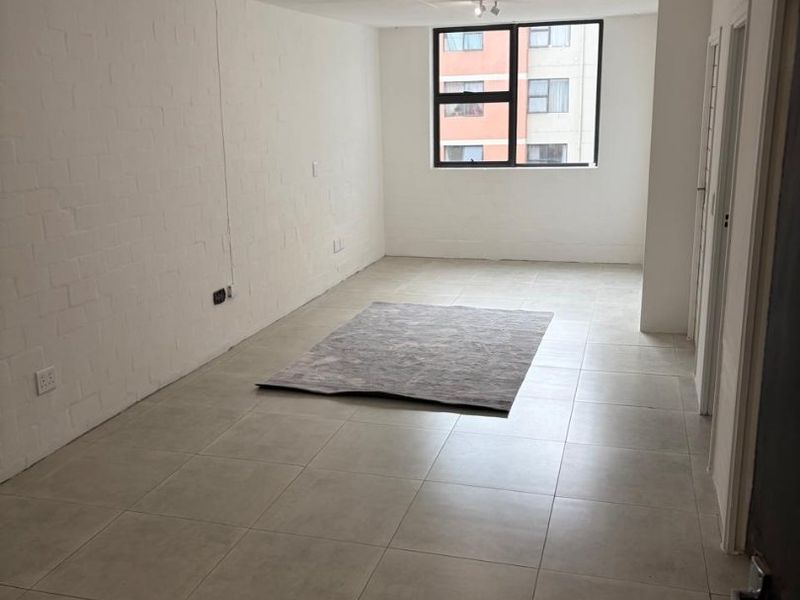 A bright, open-plan living area featuring neutral tiled flooring, painted brick walls, and large windows providing natural light.