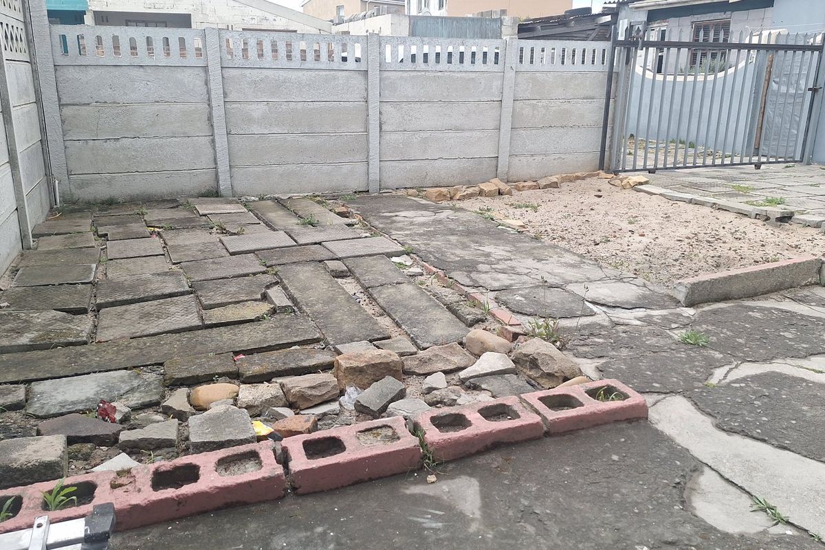 A rear outdoor area featuring a precast concrete boundary wall, metal gate, and a mix of uneven paving and concrete surfaces requiring maintenance.