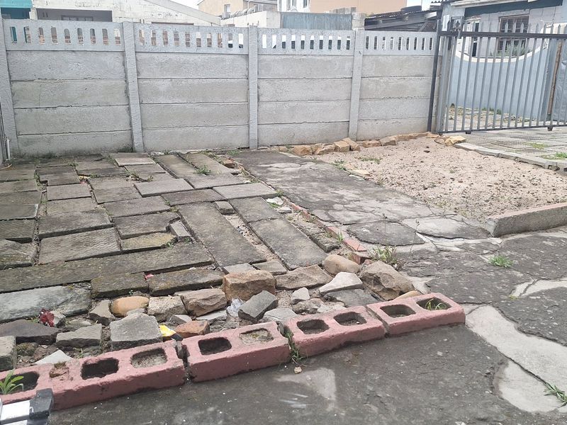 A rear outdoor area featuring a precast concrete boundary wall, metal gate, and a mix of uneven paving and concrete surfaces requiring maintenance.