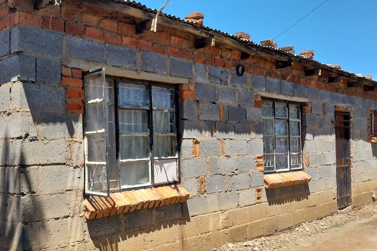 Exterior view of a single-story house featuring unfinished concrete block walls, corrugated metal roofing, and steel-framed windows with security bars.