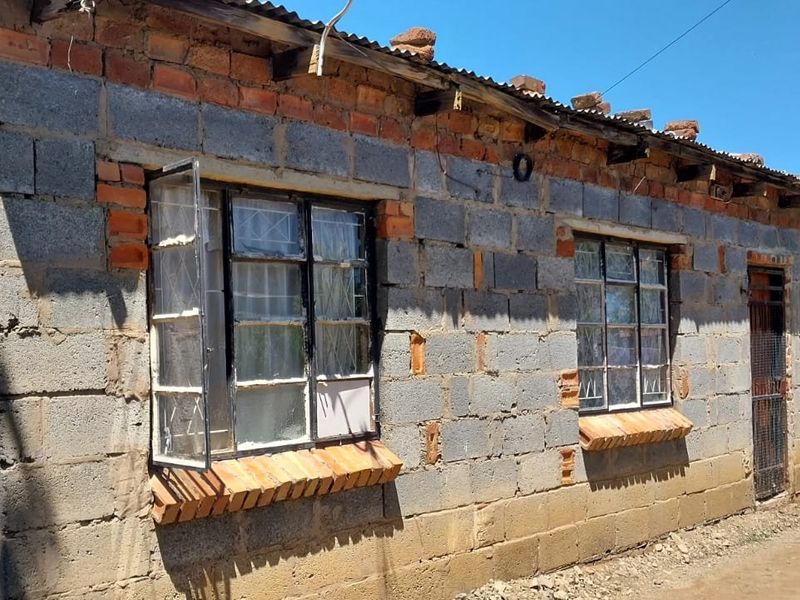 Exterior view of a single-story house featuring unfinished concrete block walls, corrugated metal roofing, and steel-framed windows with security bars.