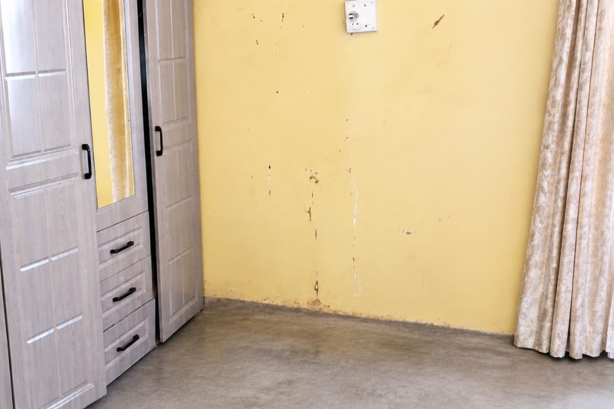 A bedroom featuring a built-in wardrobe and a window with curtains, showing visible wear and damage to the wall surfaces.