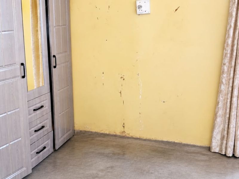A bedroom featuring a built-in wardrobe and a window with curtains, showing visible wear and damage to the wall surfaces.