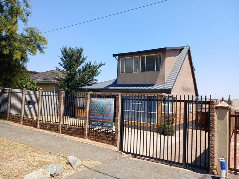 Front exterior view of a two-story house featuring a paved driveway, metal security fencing, and a brick boundary wall.