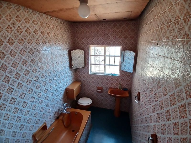 A dated bathroom featuring original colored sanitaryware, full-height patterned wall tiles, and a window with security bars.