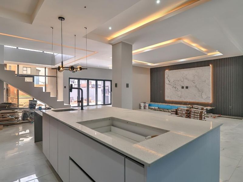 Spacious open-plan living area featuring a modern kitchen island, marble feature wall, recessed lighting, and contemporary staircase, currently in the final stages of construction.