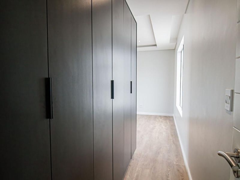 A modern, clean hallway featuring sleek dark built-in cupboards, recessed ceiling lighting, and light wood-look laminate flooring.