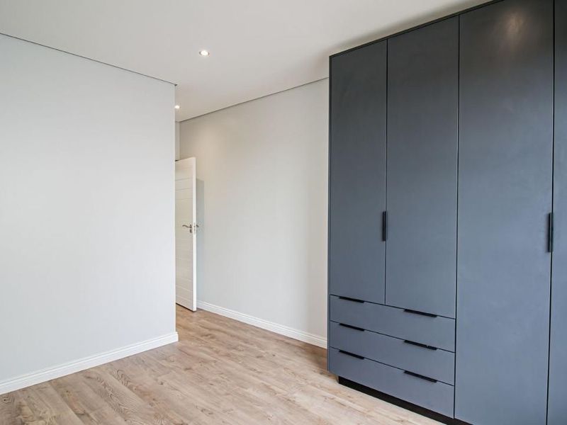 A bright, modern bedroom featuring sleek dark grey built-in cupboards, light laminate flooring, and clean neutral walls.
