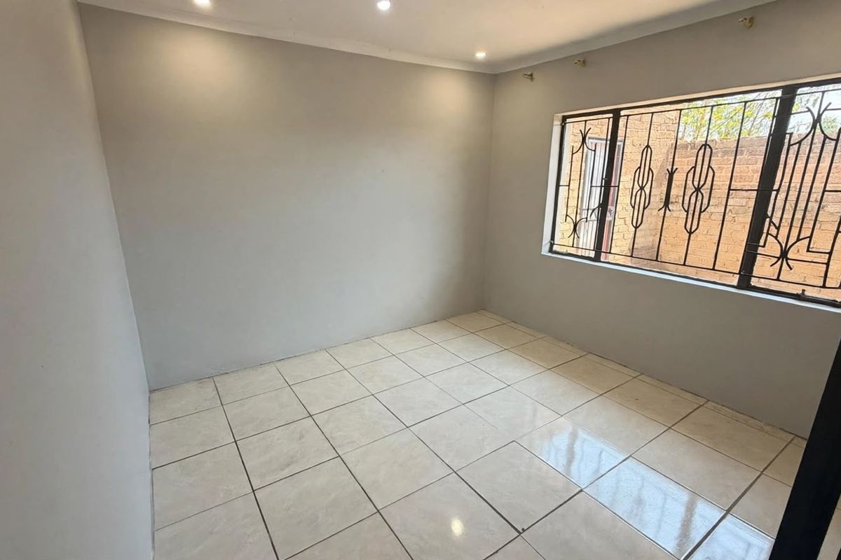 A clean, empty bedroom featuring neutral tiled flooring, recessed lighting, and a window fitted with security bars.