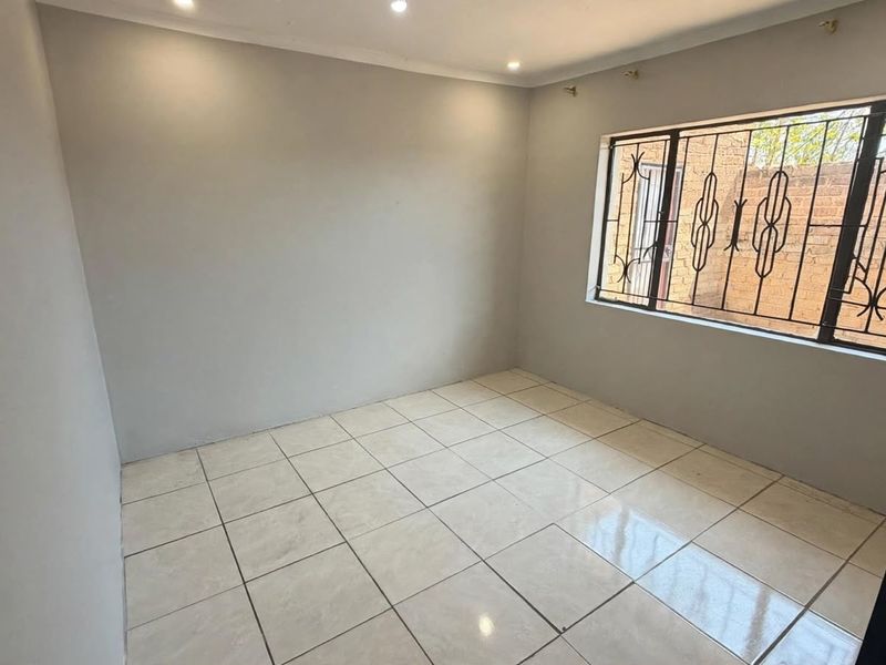 A clean, empty bedroom featuring neutral tiled flooring, recessed lighting, and a window fitted with security bars.
