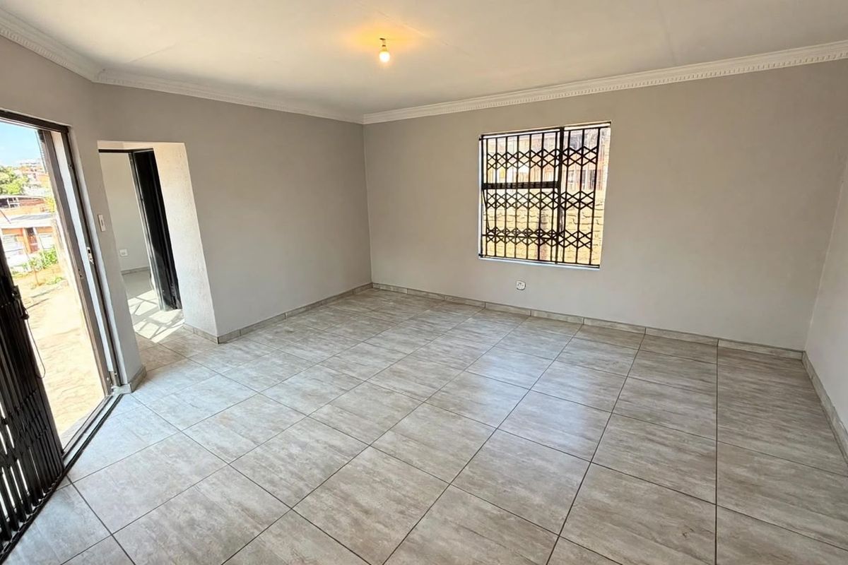 A bright, empty living room featuring neutral tiled flooring, cornice detailing, and secure access via a sliding door and window security bars.