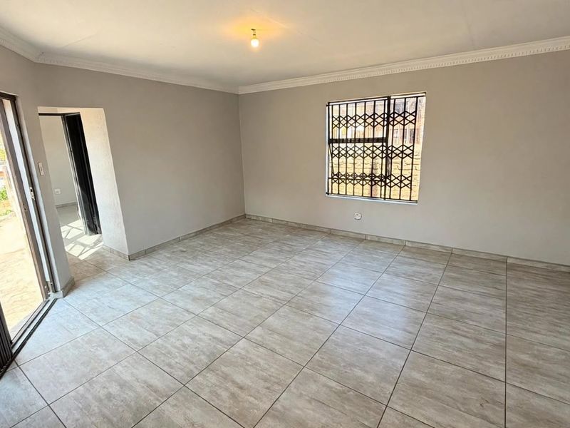 A bright, empty living room featuring neutral tiled flooring, cornice detailing, and secure access via a sliding door and window security bars.