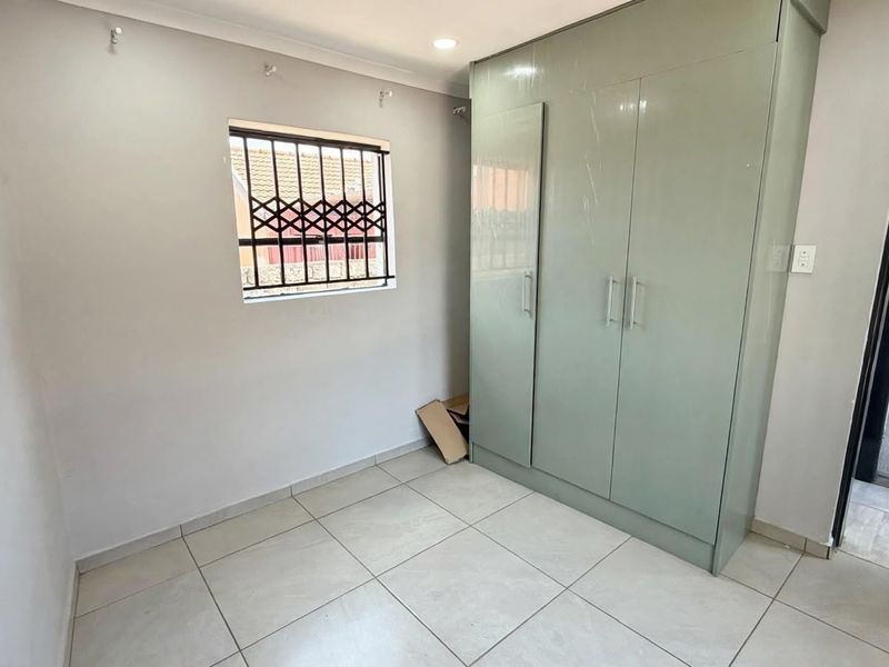A clean, empty bedroom featuring neutral tiled flooring, a built-in wardrobe, and a window fitted with security bars.