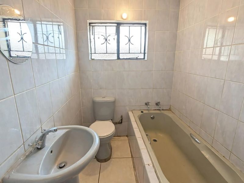 A functional bathroom featuring a pedestal basin, toilet, and built-in bathtub with full-height wall tiling and security bars on the window.