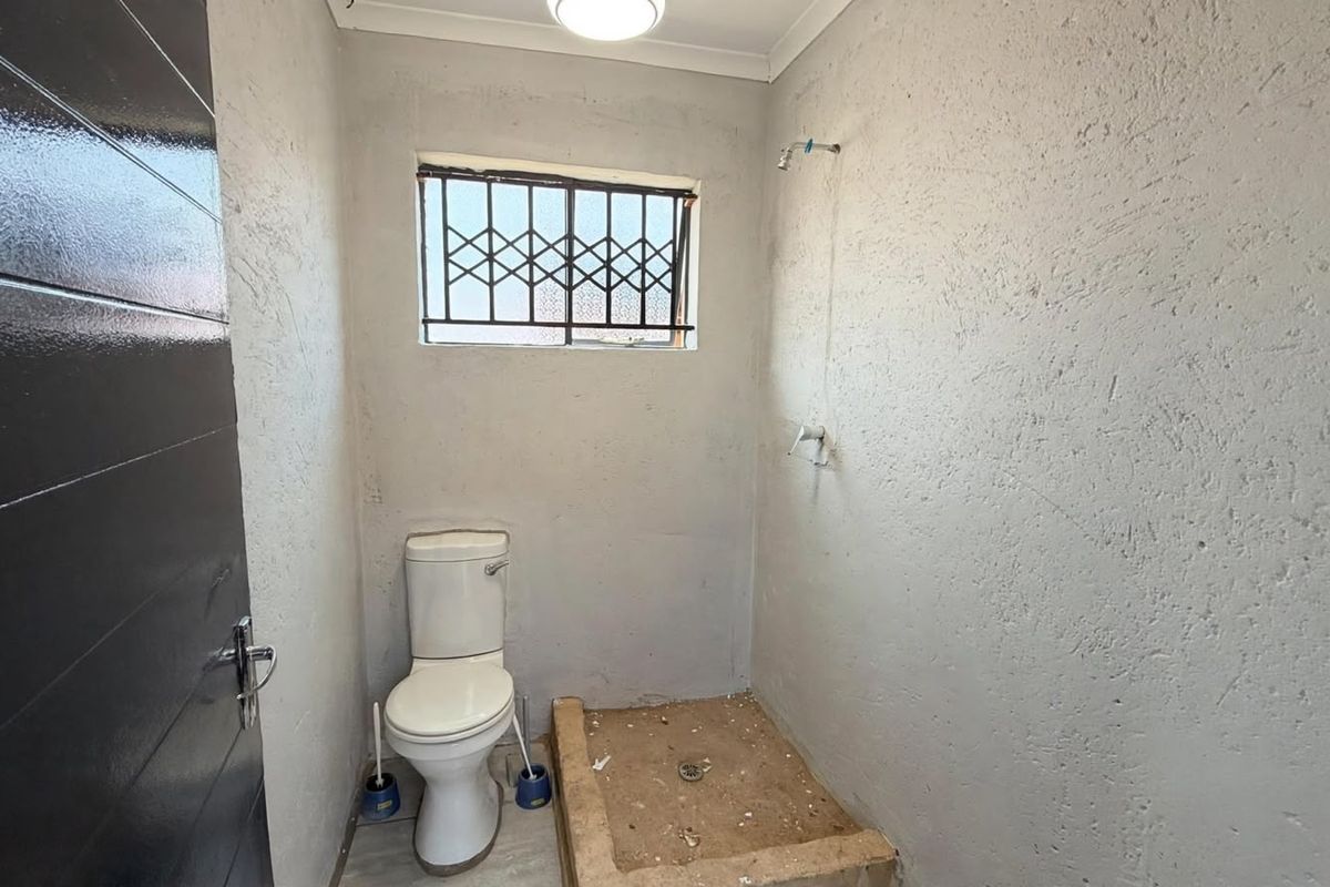 A compact bathroom featuring a toilet and an unfinished shower area with exposed wall textures and security window bars.
