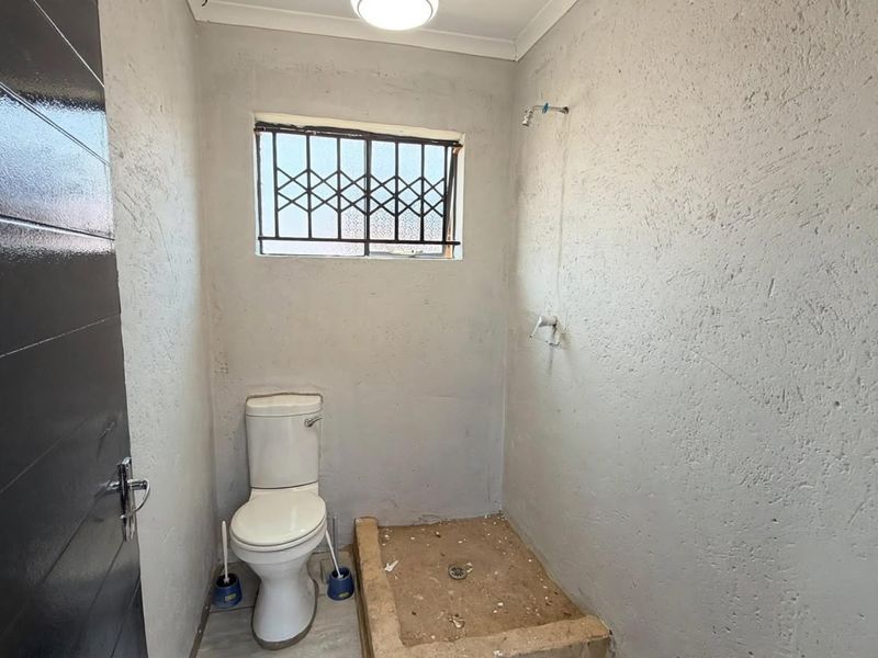 A compact bathroom featuring a toilet and an unfinished shower area with exposed wall textures and security window bars.