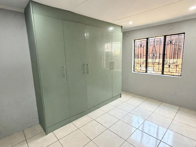 A clean, empty bedroom featuring modern sage green built-in cupboards, tiled flooring, and a window fitted with security bars.