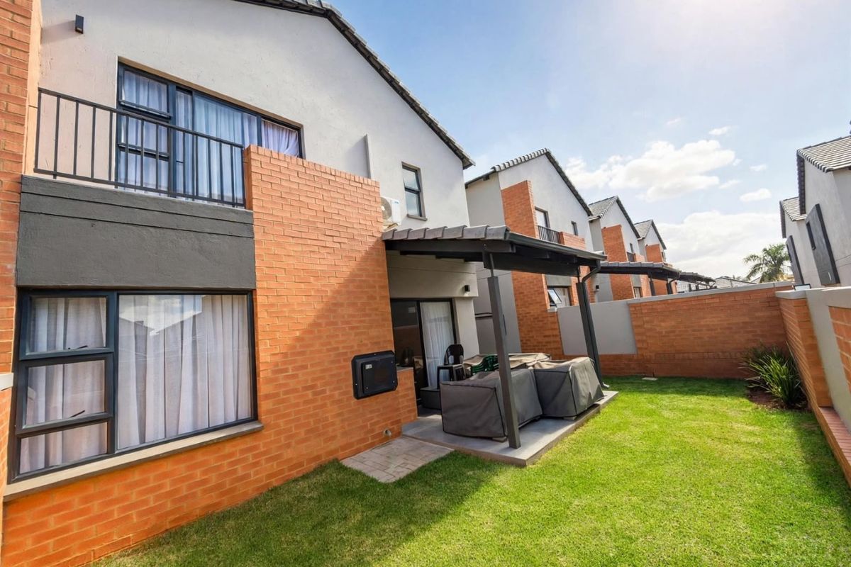 Rear view of a modern townhouse featuring a brick facade, covered patio area, balcony, and a well-maintained private garden.