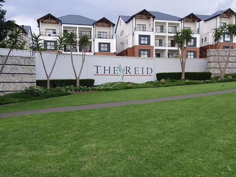Front view of The Reid apartment complex featuring a landscaped lawn, paved walkway, and modern multi-story residential buildings with stone-clad boundary walls.