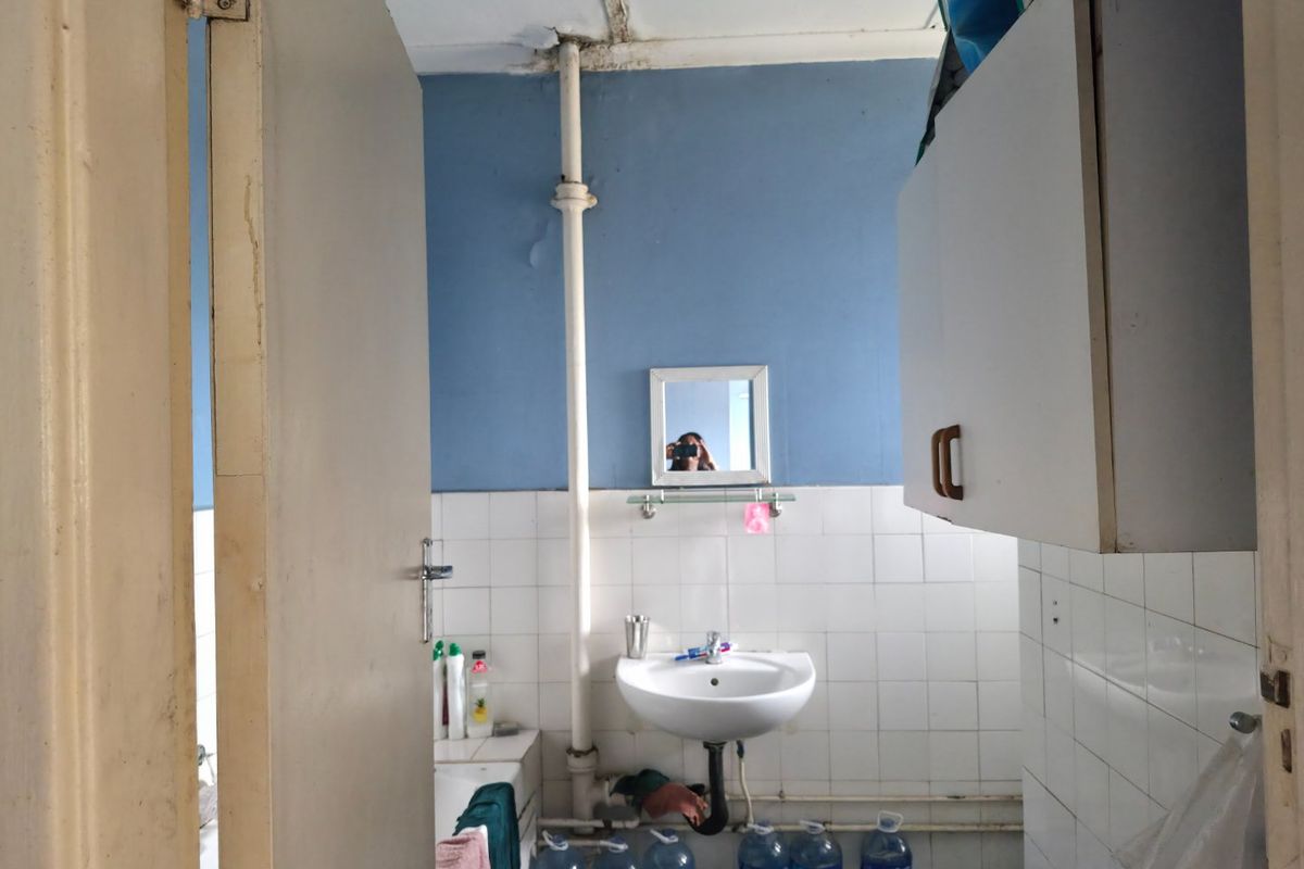 A dated bathroom featuring a pedestal washbasin, wall-mounted cabinetry, and exposed plumbing, requiring significant maintenance and cosmetic updates to improve its condition.