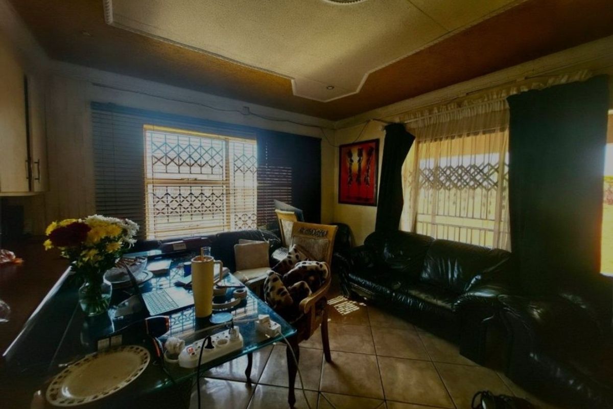 A living room featuring tiled flooring, decorative ceiling details, and security burglar bars on the windows, offering a dated space ready for modernization.
