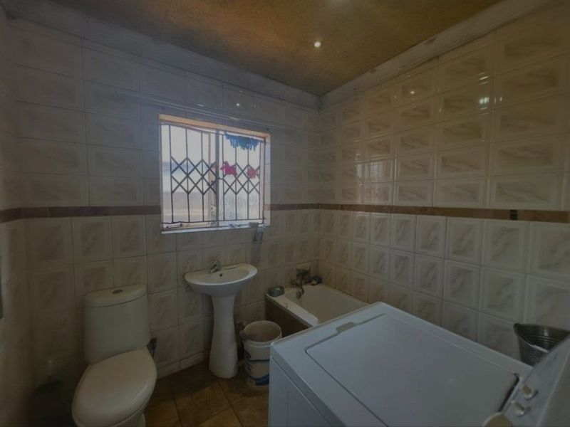 A dated bathroom featuring a pedestal basin, toilet, and bathtub with full-height wall tiling and a security gate on the window.