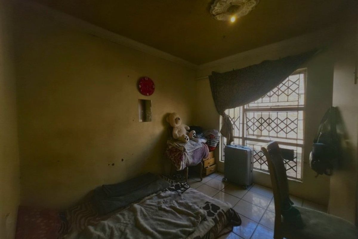 A bedroom featuring tiled flooring, security bars on the window, and a decorative ceiling rose, currently requiring cosmetic updates and wall repairs.