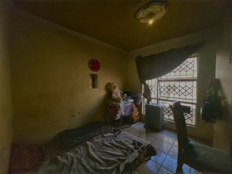 A bedroom featuring tiled flooring, security bars on the window, and a decorative ceiling rose, currently requiring cosmetic updates and wall repairs.