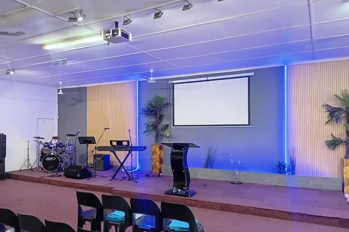 A commercial event space or clubhouse featuring a raised stage, projector screen, integrated LED accent lighting, and professional track lighting systems.