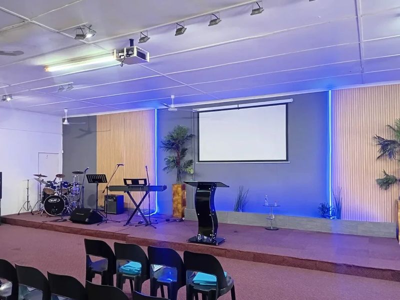 A commercial event space or clubhouse featuring a raised stage, projector screen, integrated LED accent lighting, and professional track lighting systems.