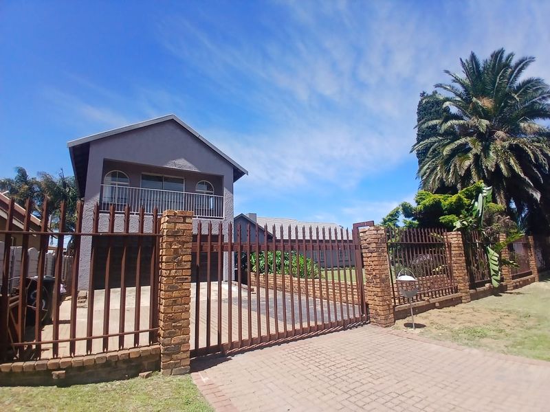 Front exterior view of a two-story house featuring a paved driveway, brick perimeter wall, and metal security fencing.