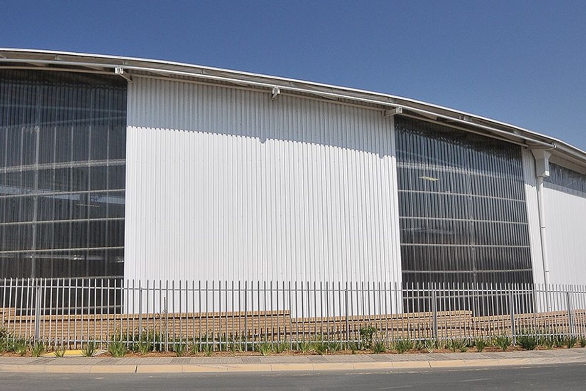 Modern industrial warehouse facility featuring a curved roof design, corrugated metal cladding, and large translucent wall panels, secured by perimeter steel palisade fencing.