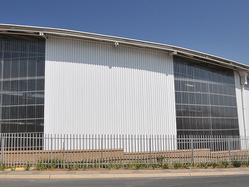 Modern industrial warehouse facility featuring a curved roof design, corrugated metal cladding, and large translucent wall panels, secured by perimeter steel palisade fencing.