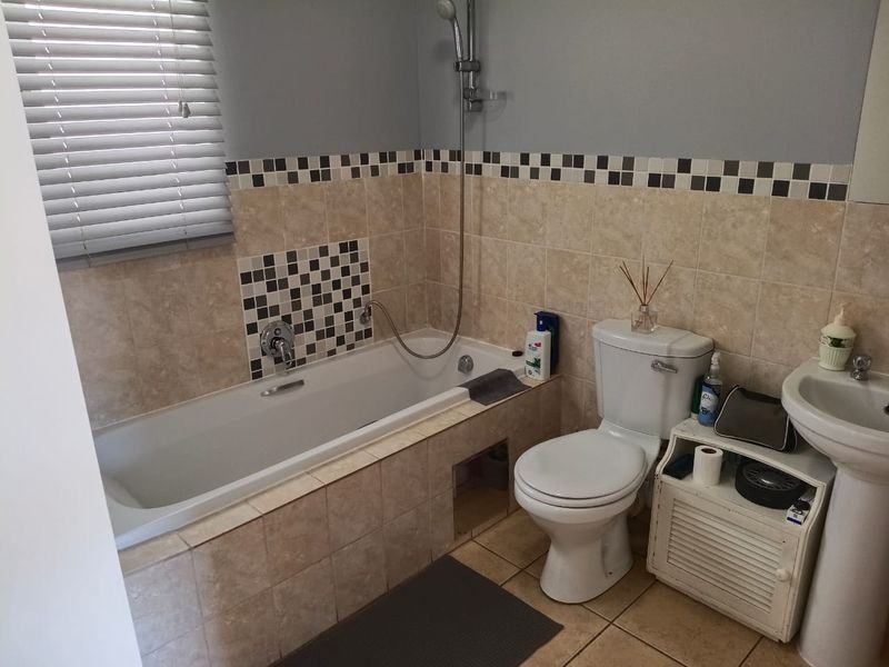 A functional bathroom featuring a built-in bathtub with shower mixer, pedestal basin, and toilet, finished with neutral wall tiles and a decorative mosaic border.