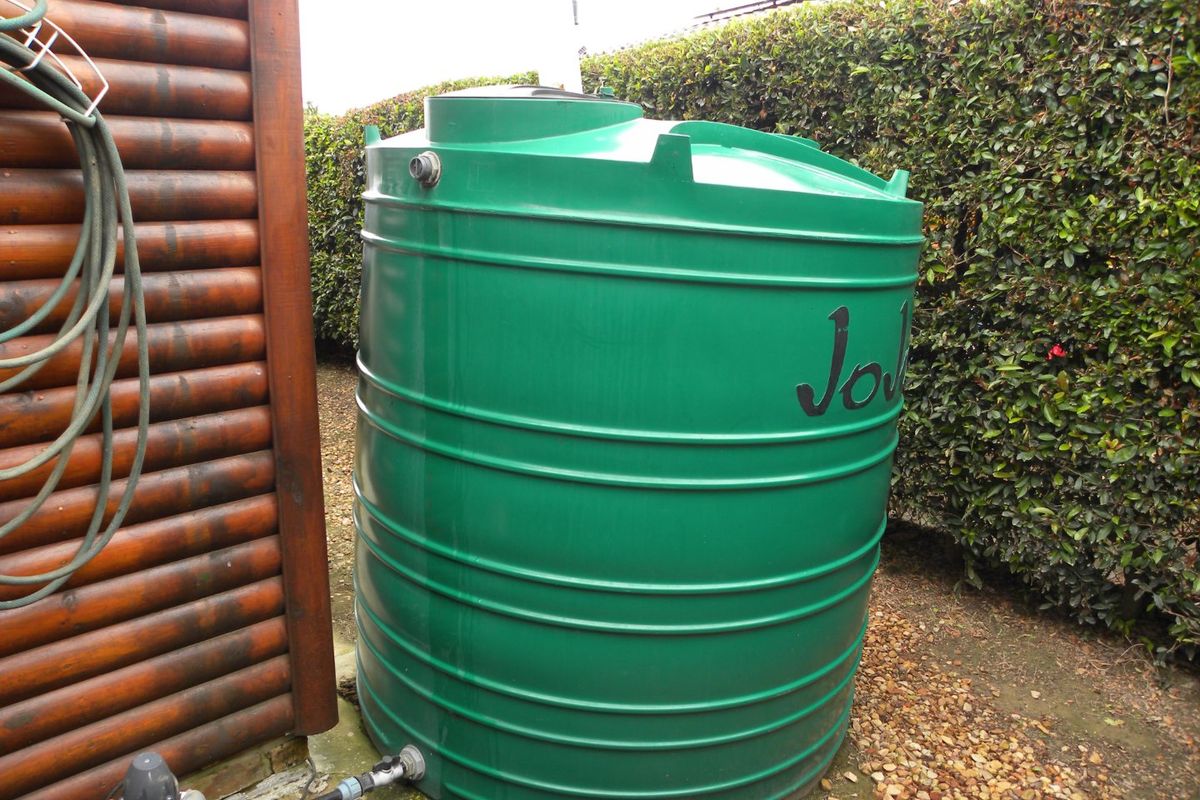A green plastic water storage tank installed in the rear garden area, featuring connected plumbing and situated next to a hedge.