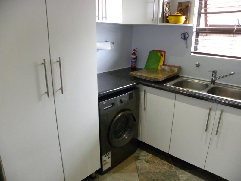 A functional scullery area featuring white built-in cabinetry, dark laminate countertops, a stainless steel double sink, and tiled flooring.