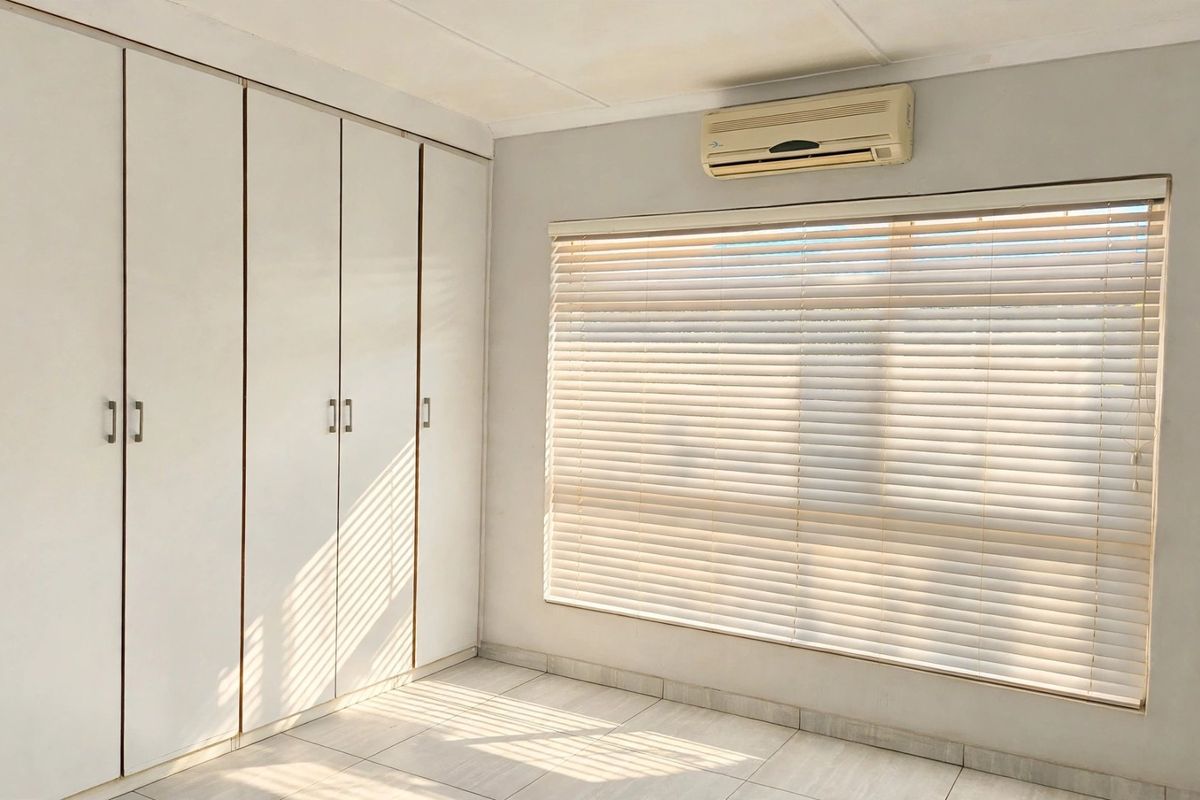 A bright, empty bedroom featuring built-in cupboards, tiled flooring, and a wall-mounted air conditioning unit with fitted venetian blinds.