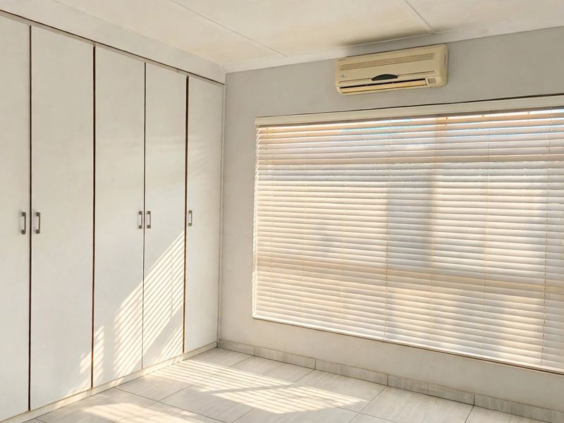 A bright, empty bedroom featuring built-in cupboards, tiled flooring, and a wall-mounted air conditioning unit with fitted venetian blinds.