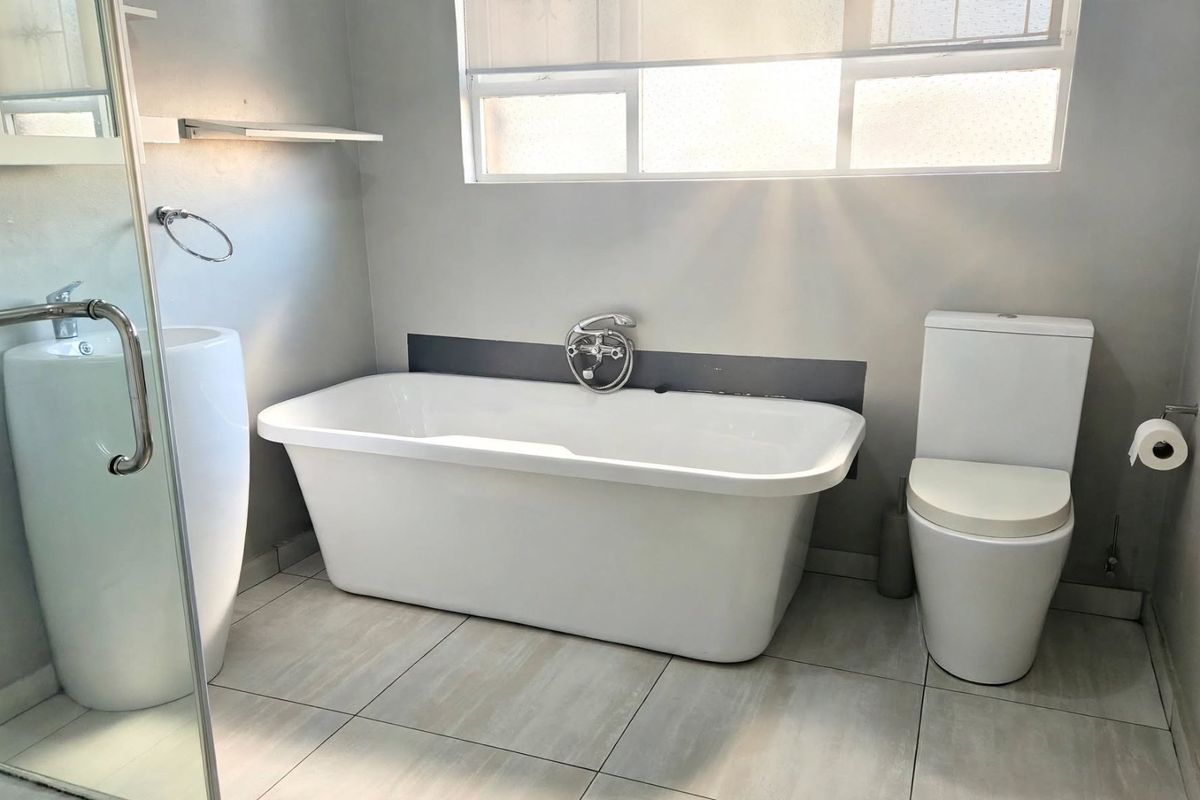 A modern, minimalist bathroom featuring a freestanding bathtub, pedestal basin, and a glass-enclosed shower with neutral floor tiling.