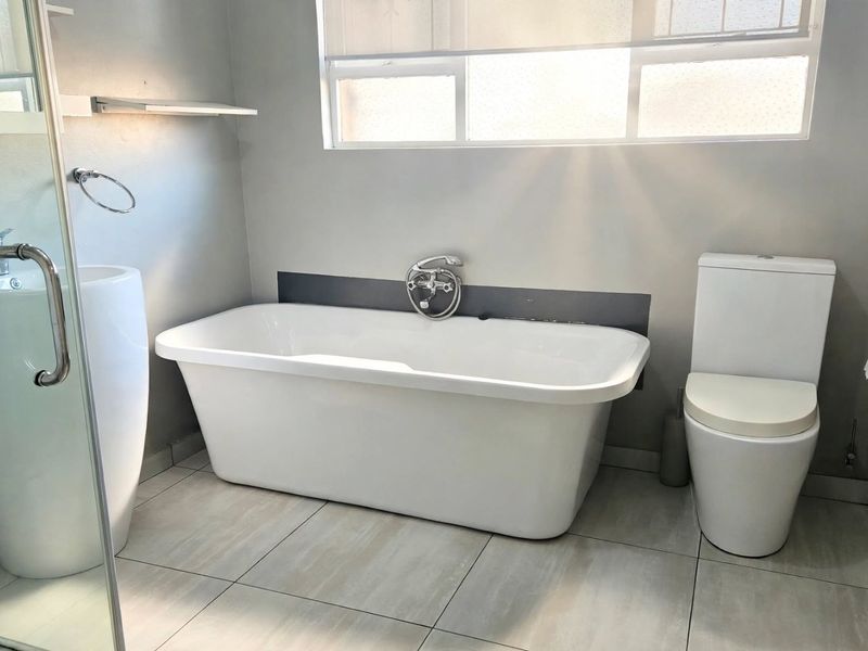 A modern, minimalist bathroom featuring a freestanding bathtub, pedestal basin, and a glass-enclosed shower with neutral floor tiling.