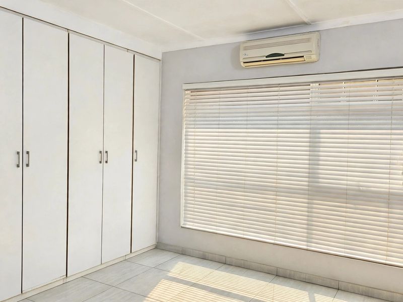 A bright, empty bedroom featuring neutral tiled flooring, ample built-in cupboard storage, and a wall-mounted air conditioning unit.