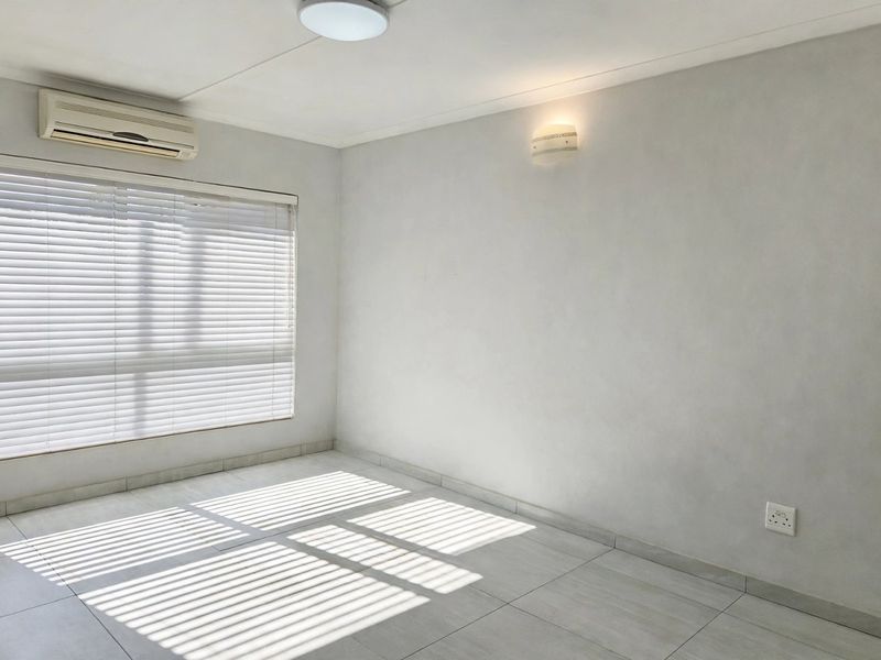 A bright, empty bedroom featuring neutral tiled flooring, an integrated air conditioning unit, and venetian blinds for adjustable natural light.