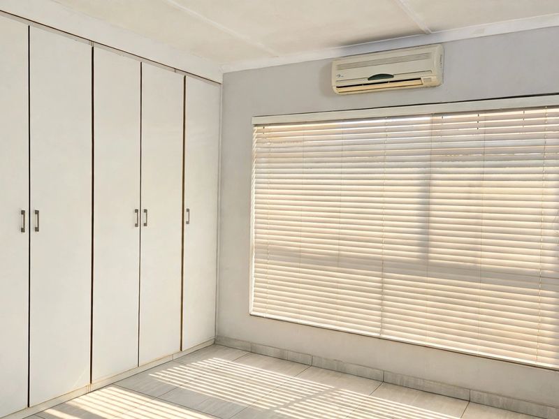 A bright, empty bedroom featuring built-in cupboards, tiled flooring, and an installed air conditioning unit with window blinds.