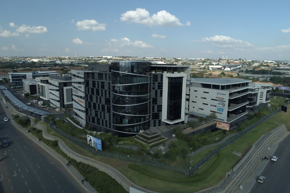 Aerial view of a modern multi-story commercial office complex featuring glass facade architecture, landscaped grounds, and secure perimeter fencing in an urban setting.