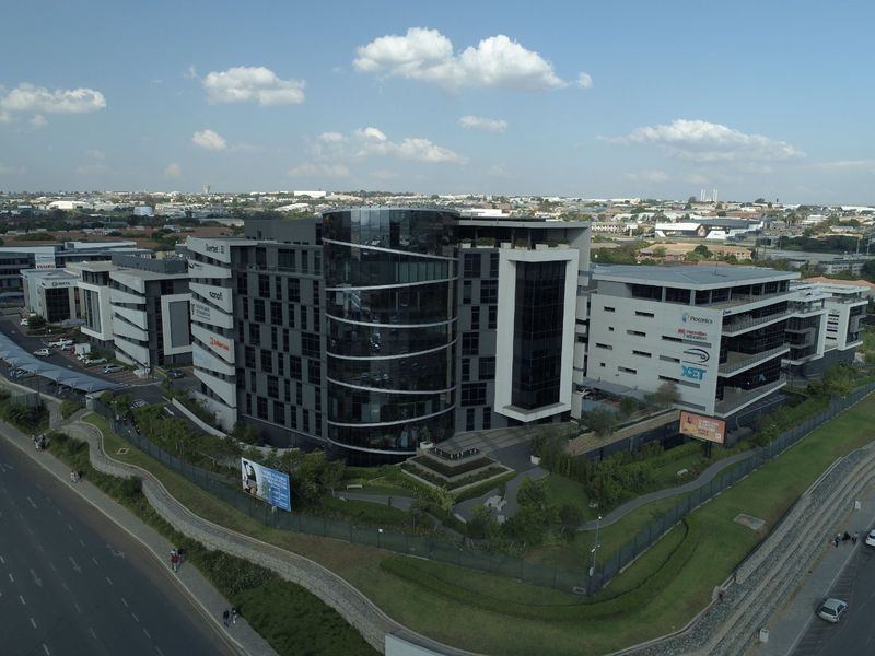 Aerial view of a modern multi-story commercial office complex featuring glass facade architecture, landscaped grounds, and secure perimeter fencing in an urban setting.